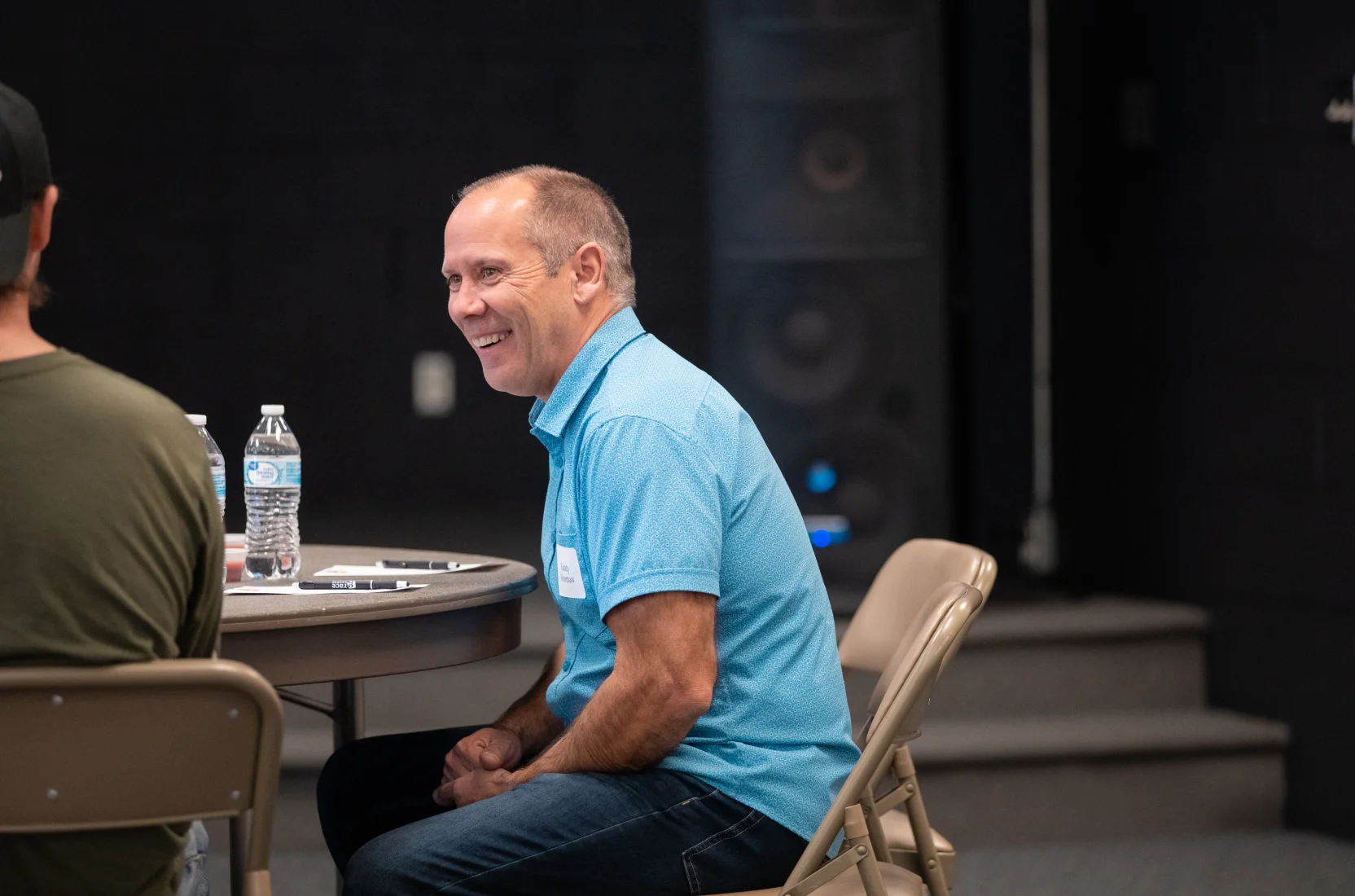 Man in a light blue shirt smiling and sitting at a round table during a small group discussion.