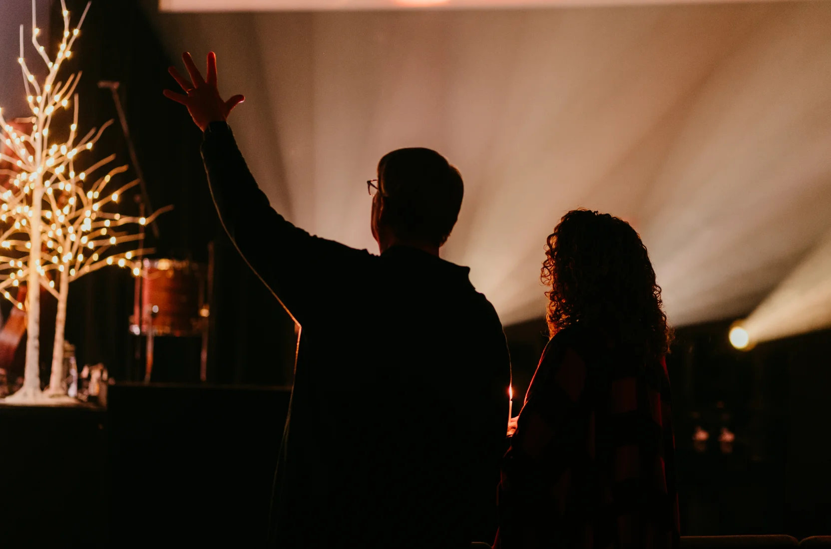 Silhouette of a man and woman worshipping with raised hands in a dimly lit indoor setting with decorative lights.