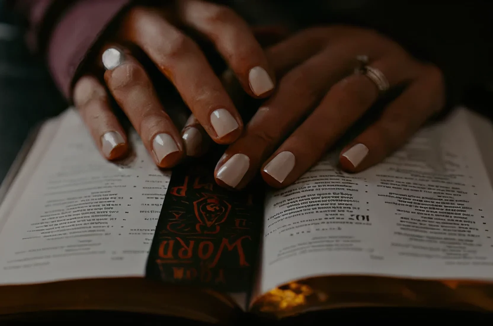 Hands with white nail polish resting on an open Bible with a black and red bookmark.