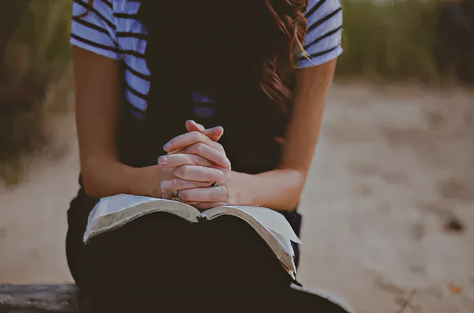 Person with clasped hands resting on an open book, sitting outdoors on a bench.