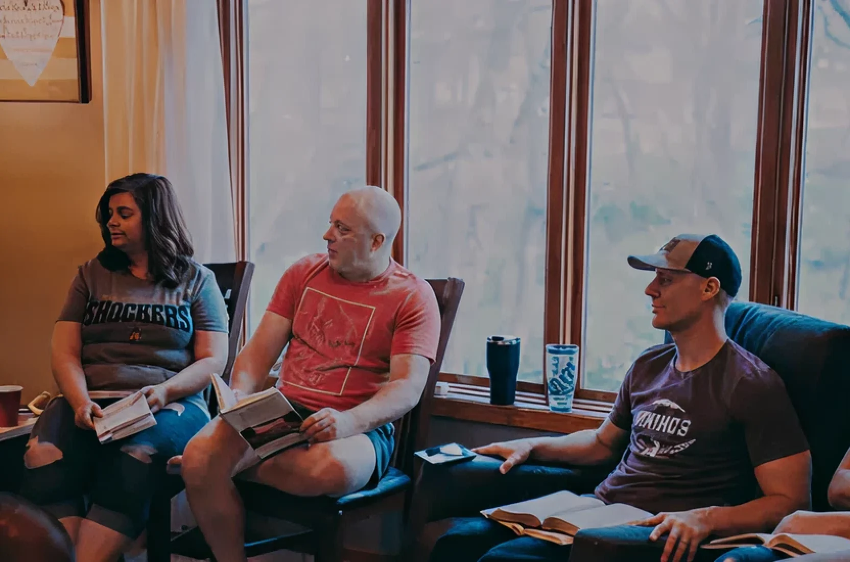 Three people sitting indoors near large windows, each holding and reading a book while engaged in conversation.