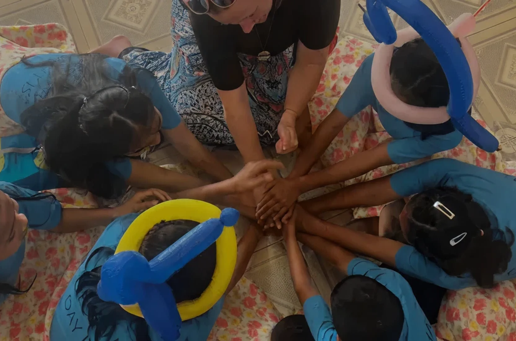 Children and an adult sitting in a circle with hands stacked together in the center, showing teamwork.