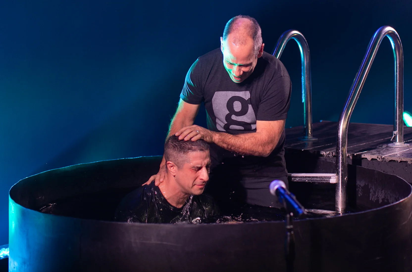 Pastor with hand on head baptizing a man in a round water tank during a service.