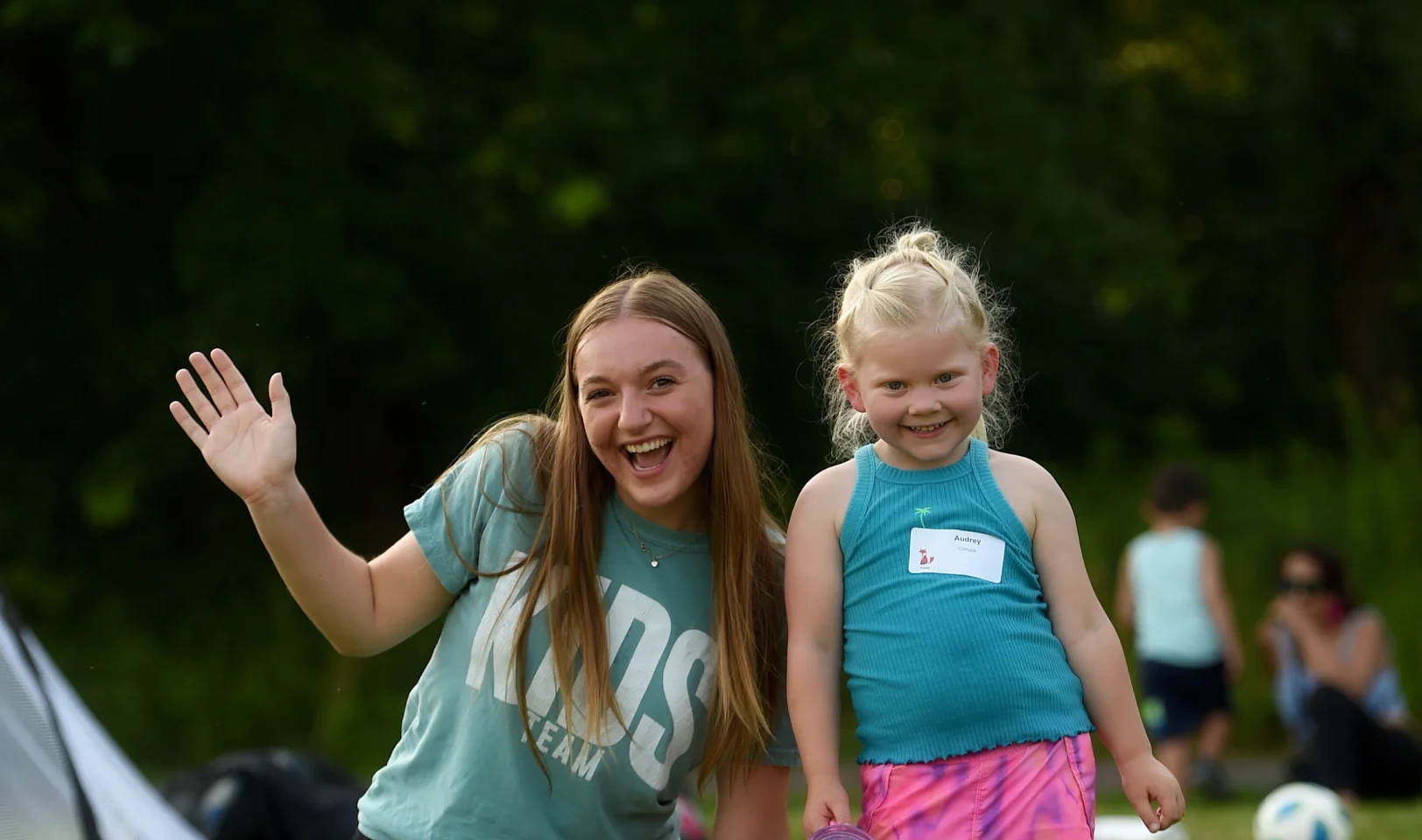 Smiling young woman waving next to a smiling young girl with a name tag outdoors.