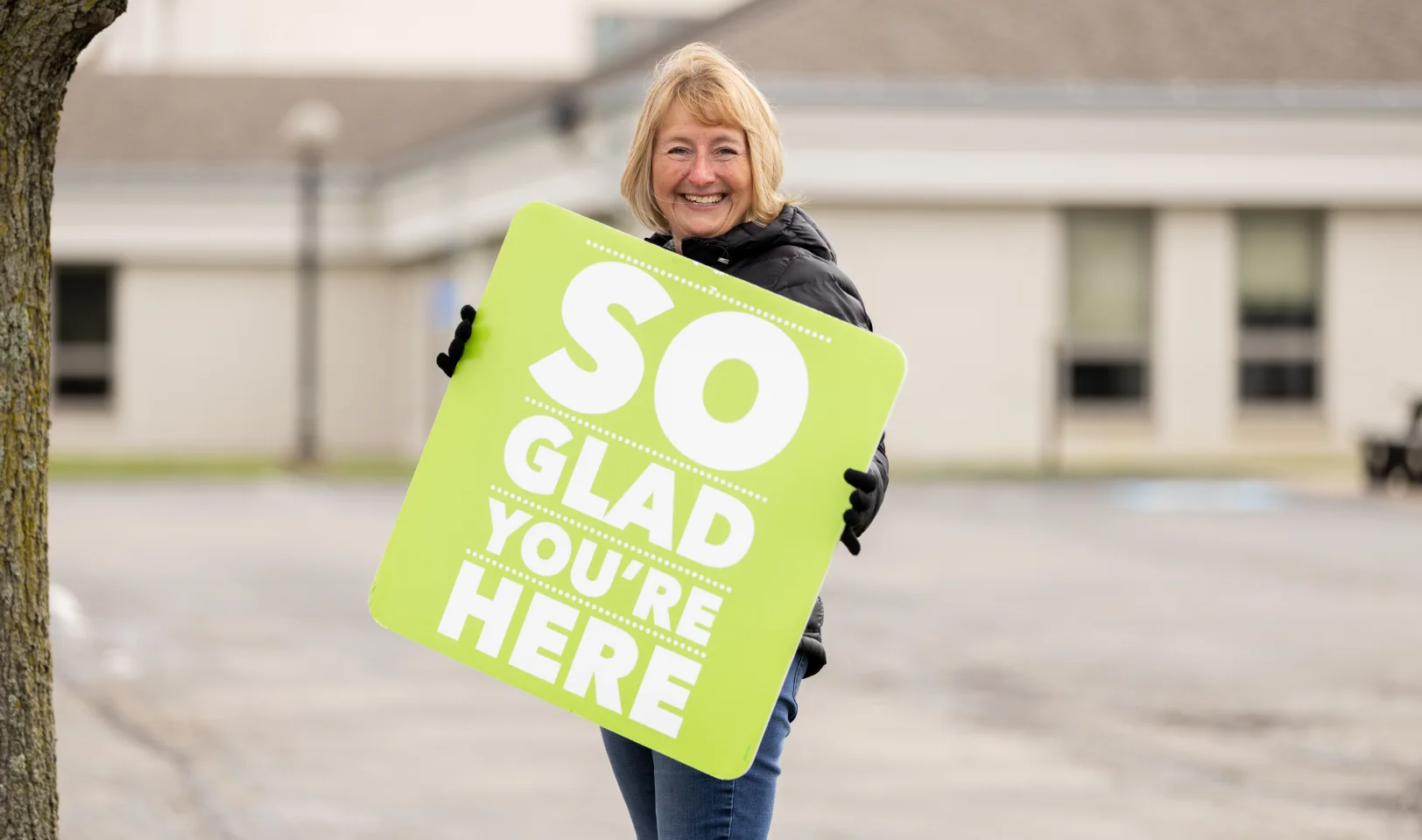 Smiling woman wearing a black jacket and gloves holding a green sign that reads, 'SO GLAD YOU'RE HERE.'