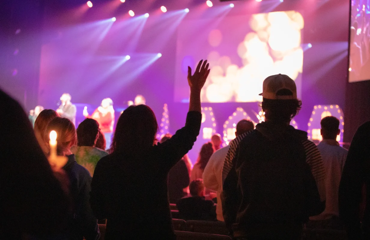 Congregation attending a worship service with hands raised and candlelight in a dimly lit auditorium with stage lights.