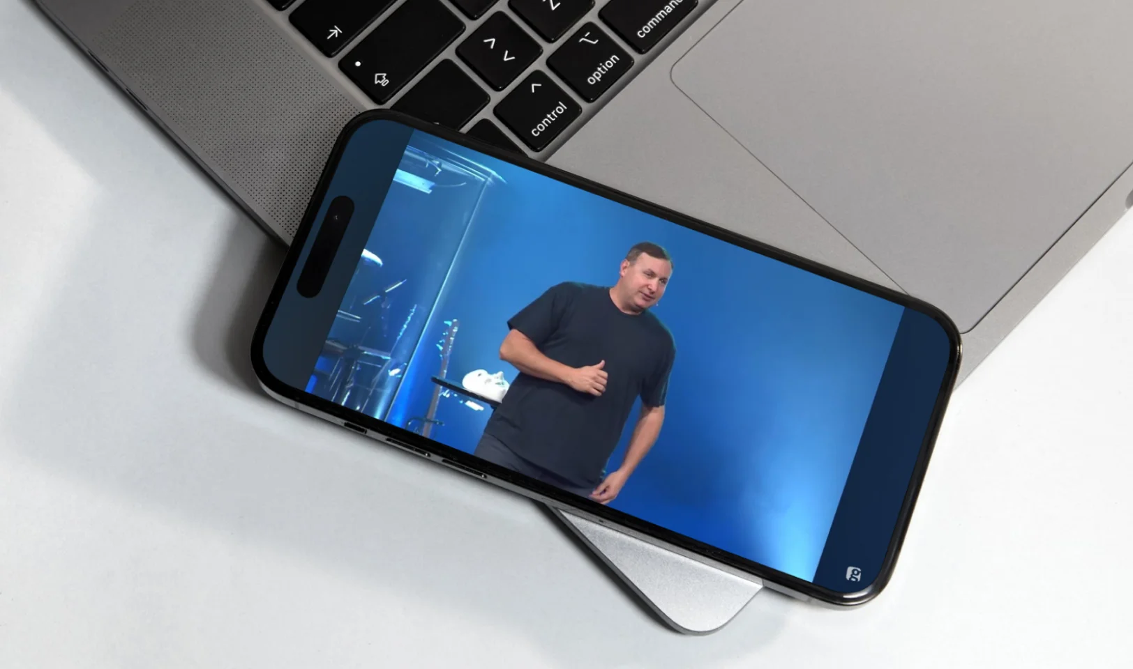 Smartphone displaying a man speaking on a blue-lit stage, placed on a silver laptop keyboard.