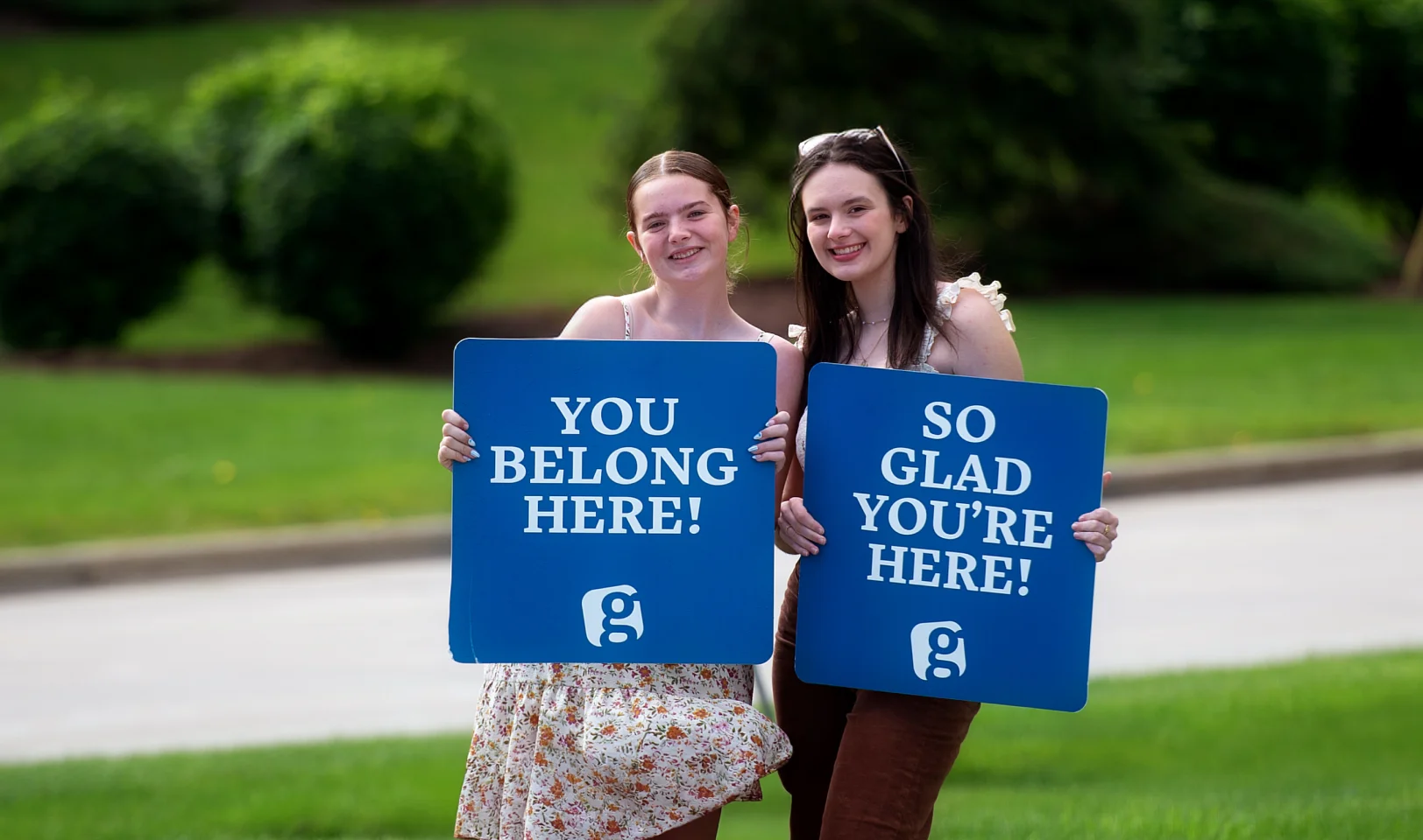 Two smiling young women outdoors holding blue signs that read 'YOU BELONG HERE!' and 'SO GLAD YOU'RE HERE!'