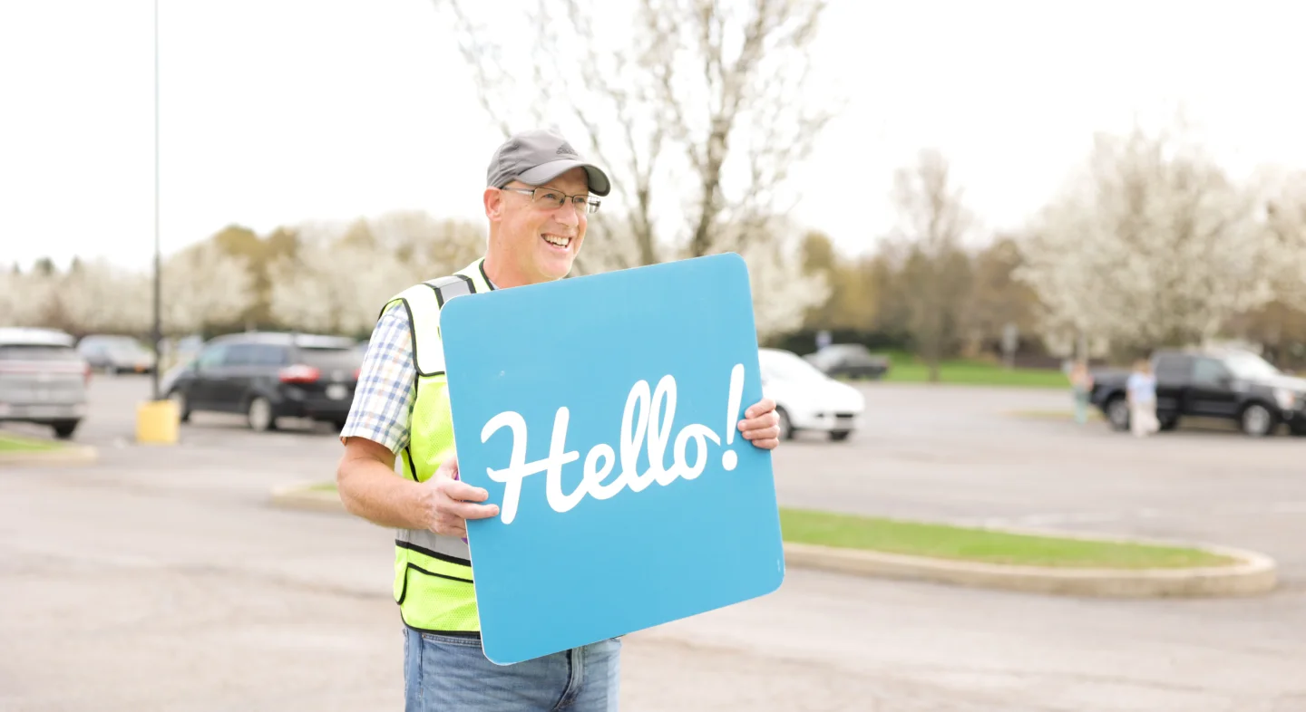 Smiling man in a safety vest and cap holding a blue sign with the word 'Hello!' in a parking lot.