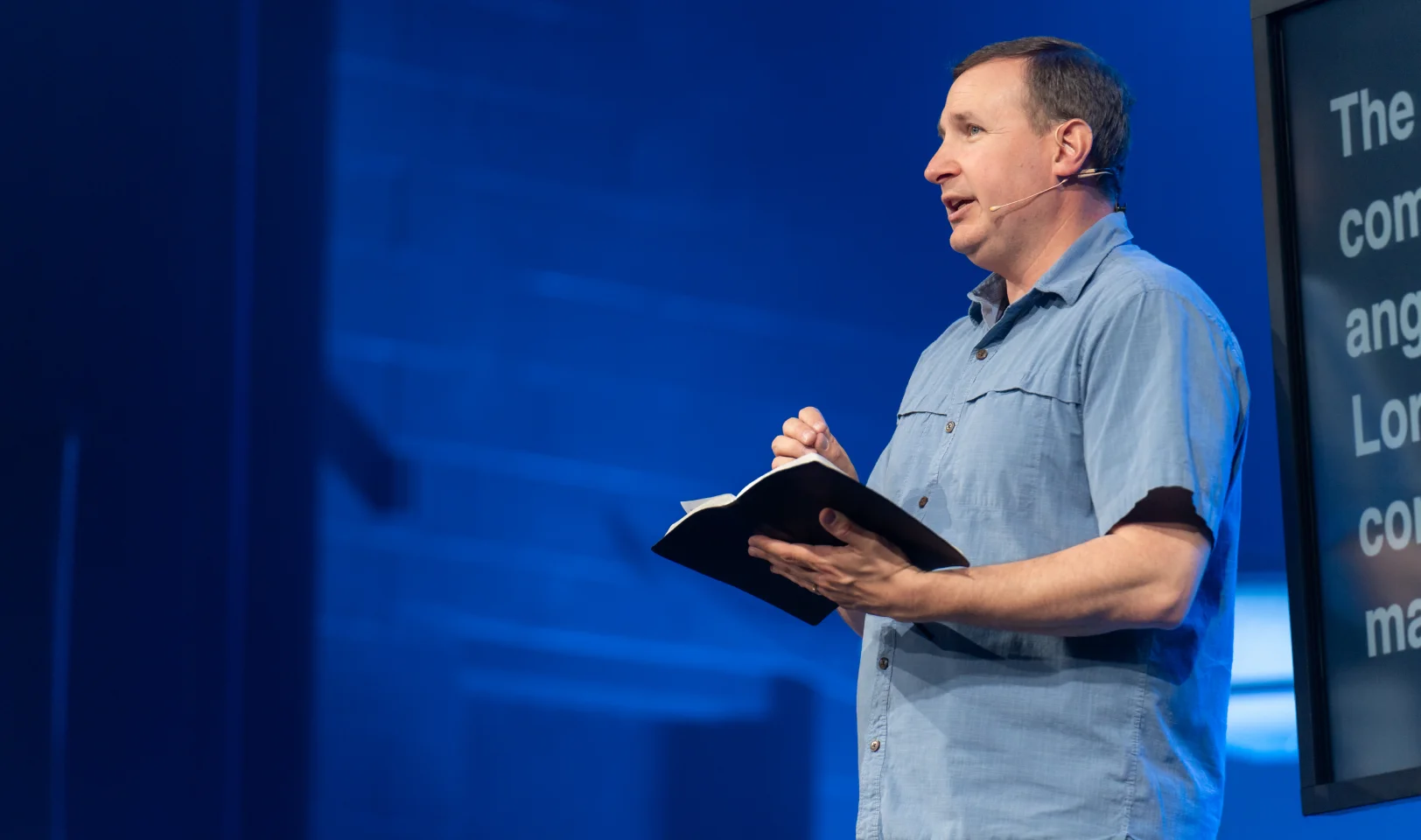 Man wearing a light blue shirt speaking on stage while holding an open book and wearing a headset microphone.
