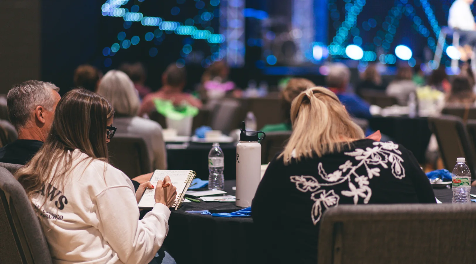 People sitting at round tables during an indoor event with stage lights in the background.