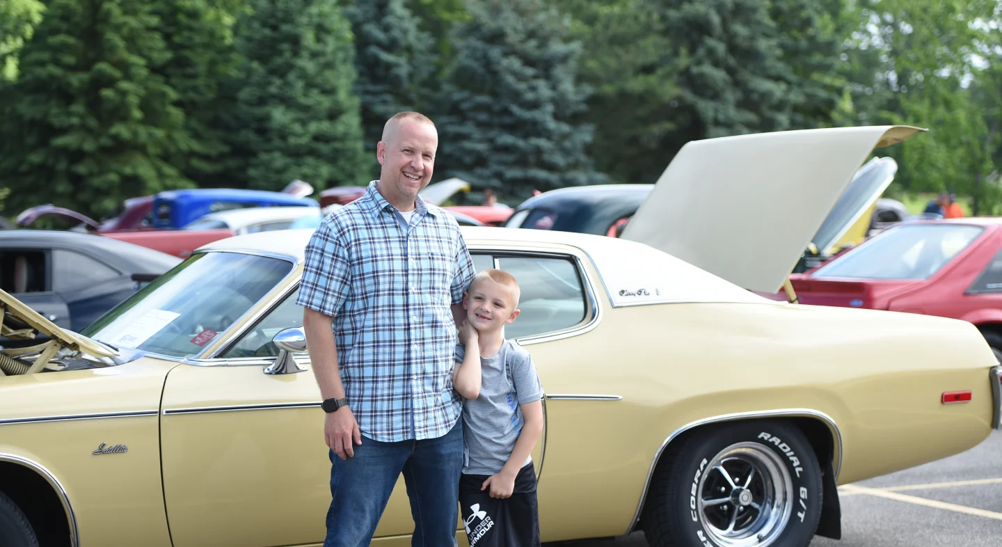Smiling father and son standing in front of a yellow classic car with its hood open at an outdoor car show.