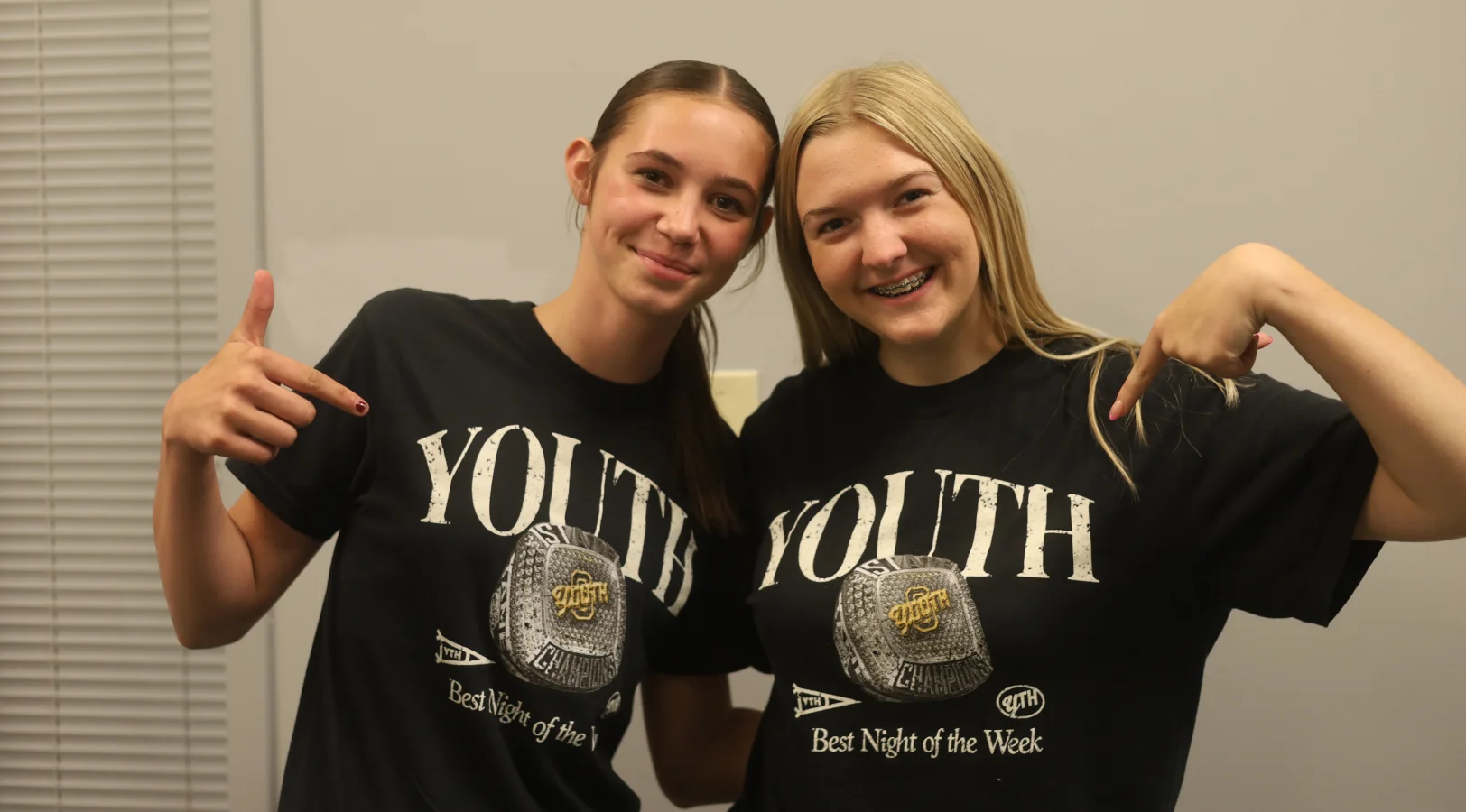 Two smiling teenage girls wearing black 'YOUTH Best Night of the Week' T-shirts, each pointing to the text on their shirts.