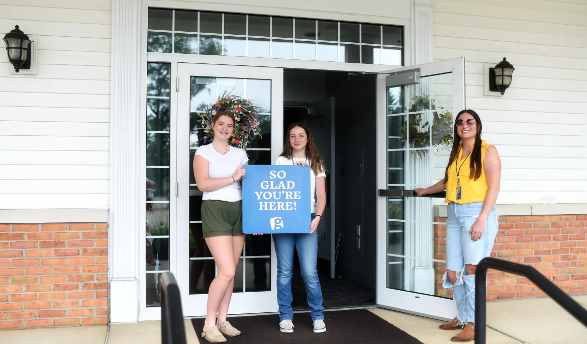 Two girls standing in front of a building entrance holding a blue sign that says 'SO GLAD YOU’RE HERE!' and a woman holding the open door.