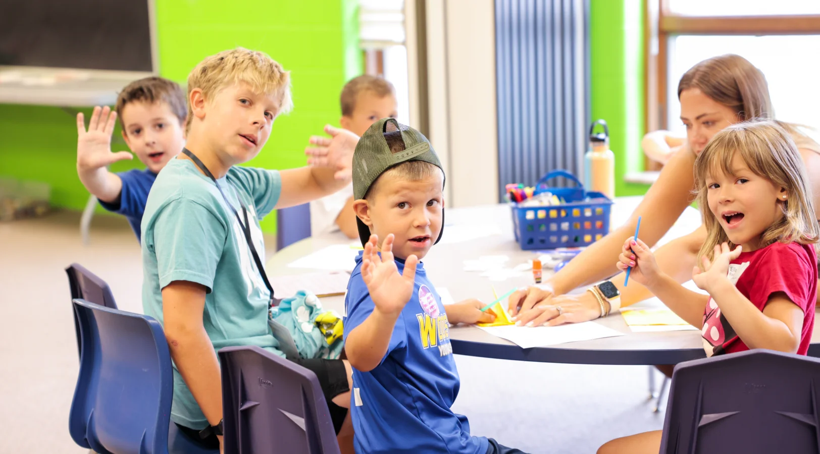 Group of children sitting at a table waving and engaging in arts and crafts with a woman assisting them.