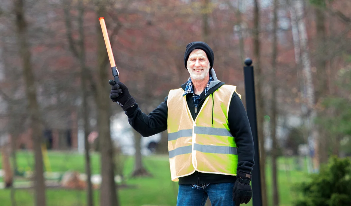 Smiling man wearing a reflective safety vest and black beanie, holding a lighted traffic baton outdoors.