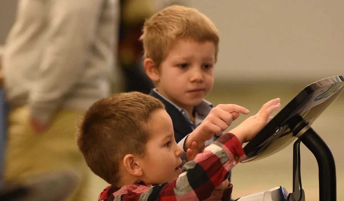 Two young boys interacting with a touchscreen kiosk or display, one pointing and the other reaching out to touch it.
