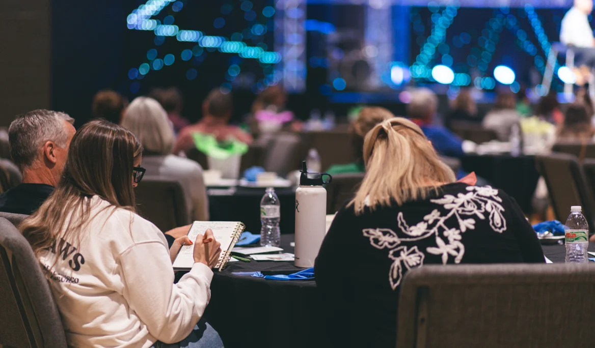 People seated at round tables attentively taking notes during a church message event.