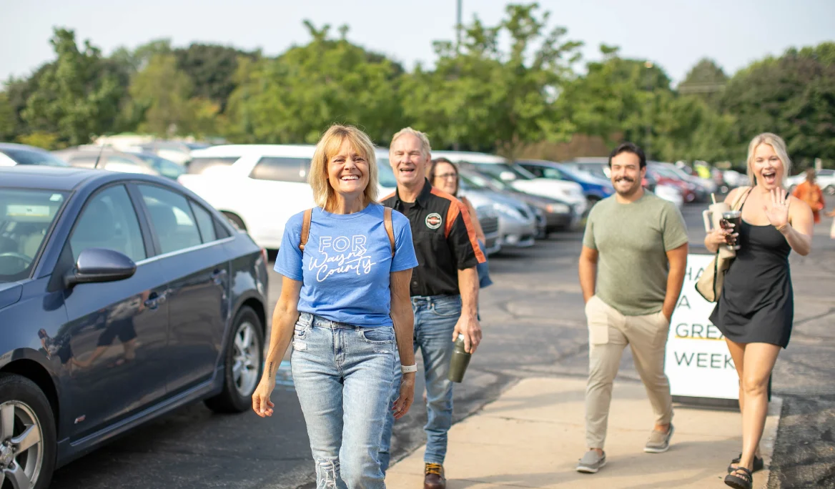 Group of four adults smiling and walking on a parking lot sidewalk with parked cars and trees in the background.