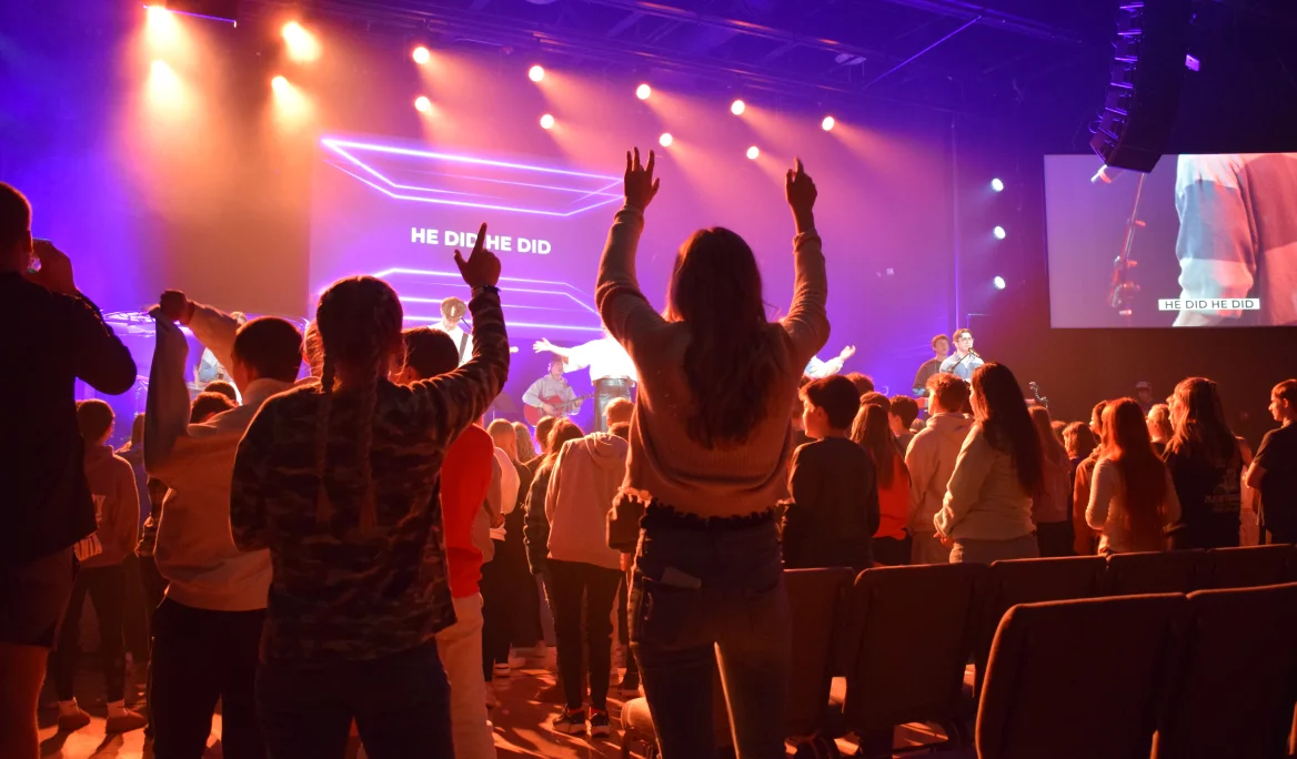 Youth worship service with raised hands, colorful stage lights, and text 'HE DID HE DID' on a screen.