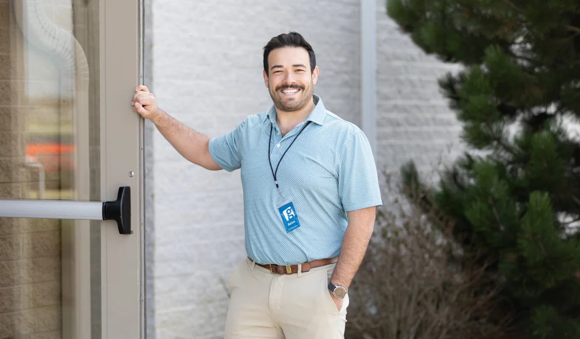 Smiling man in a blue shirt holding open a glass door outside a building.