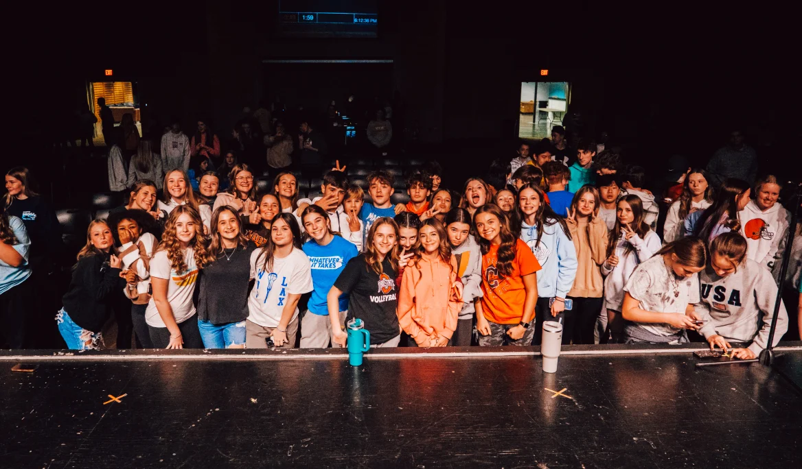 Large group of smiling teenagers posing together on a stage in a dimly lit auditorium.