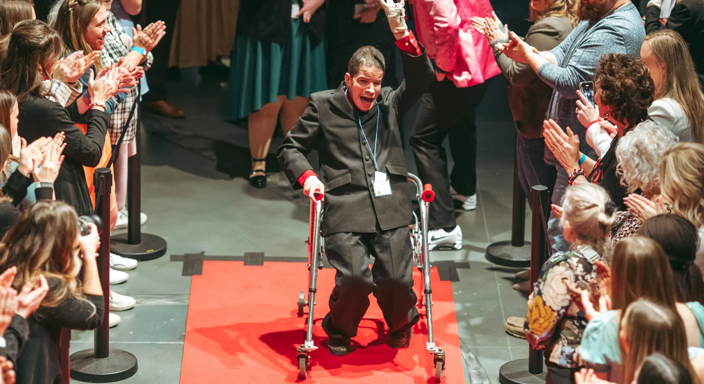 Man using a walker joyfully raising his arm while walking down a red carpet as a crowd applauds.