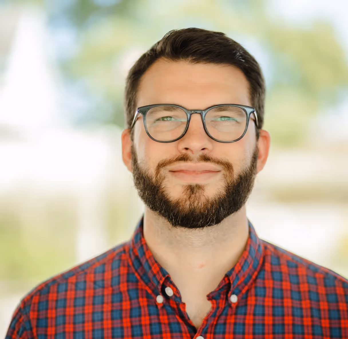 Bearded man in glasses outdoors wearing a red and blue plaid shirt.