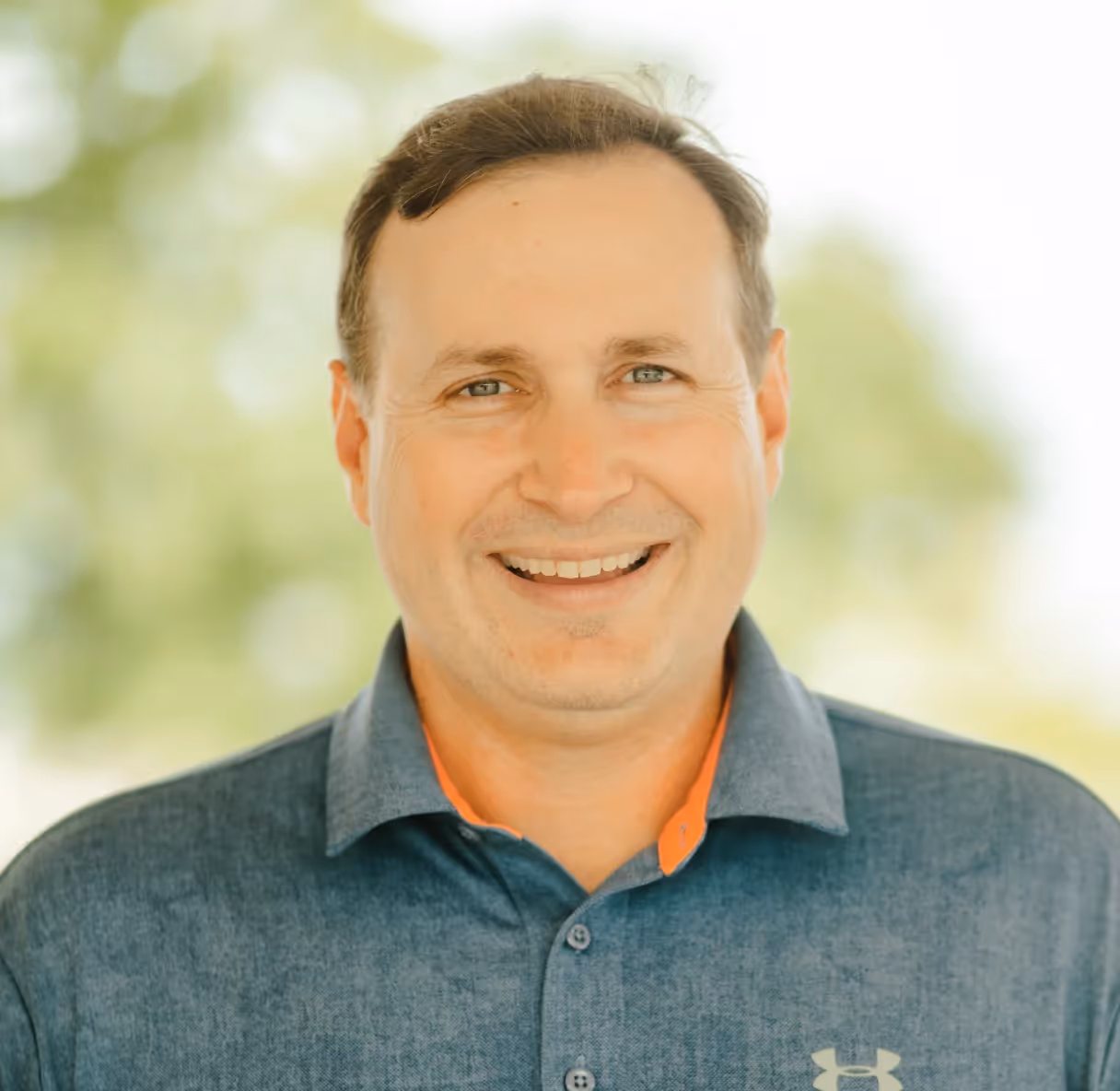 Smiling man outdoors wearing a blue collared Under Armour shirt.