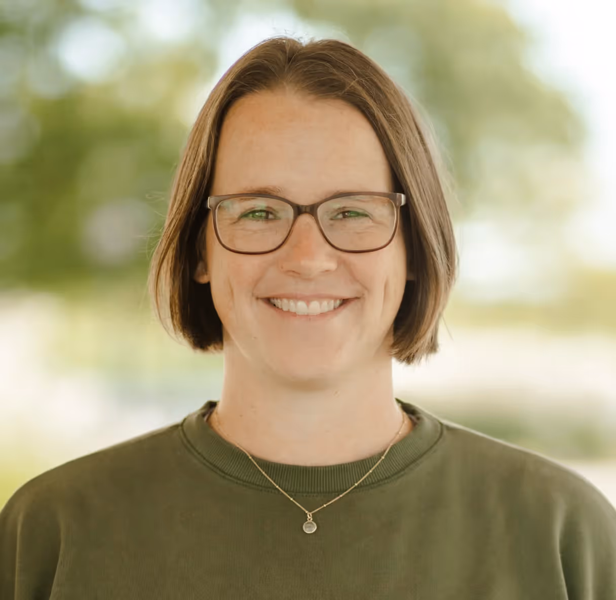 Smiling woman with short brown hair and glasses outdoors wearing a green sweatshirt.