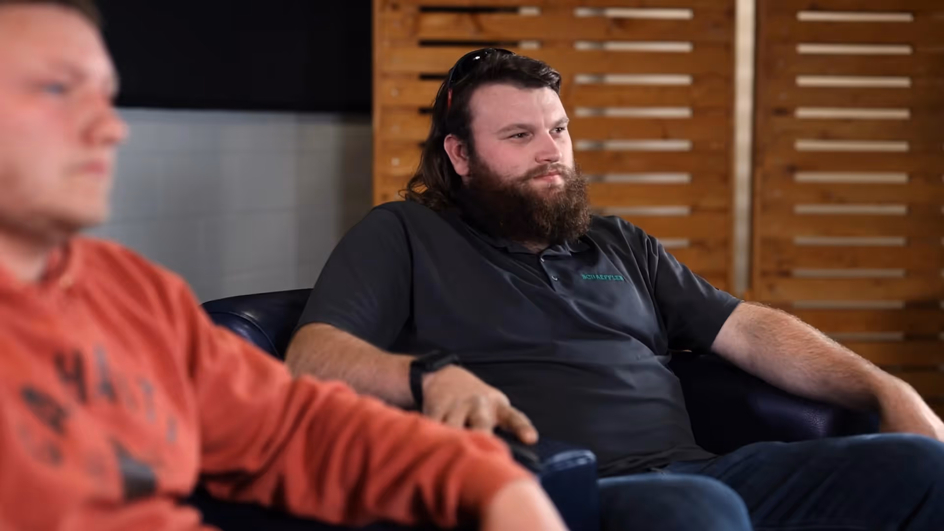 Two men seated indoors in discussion, one wearing a red shirt, one in black.