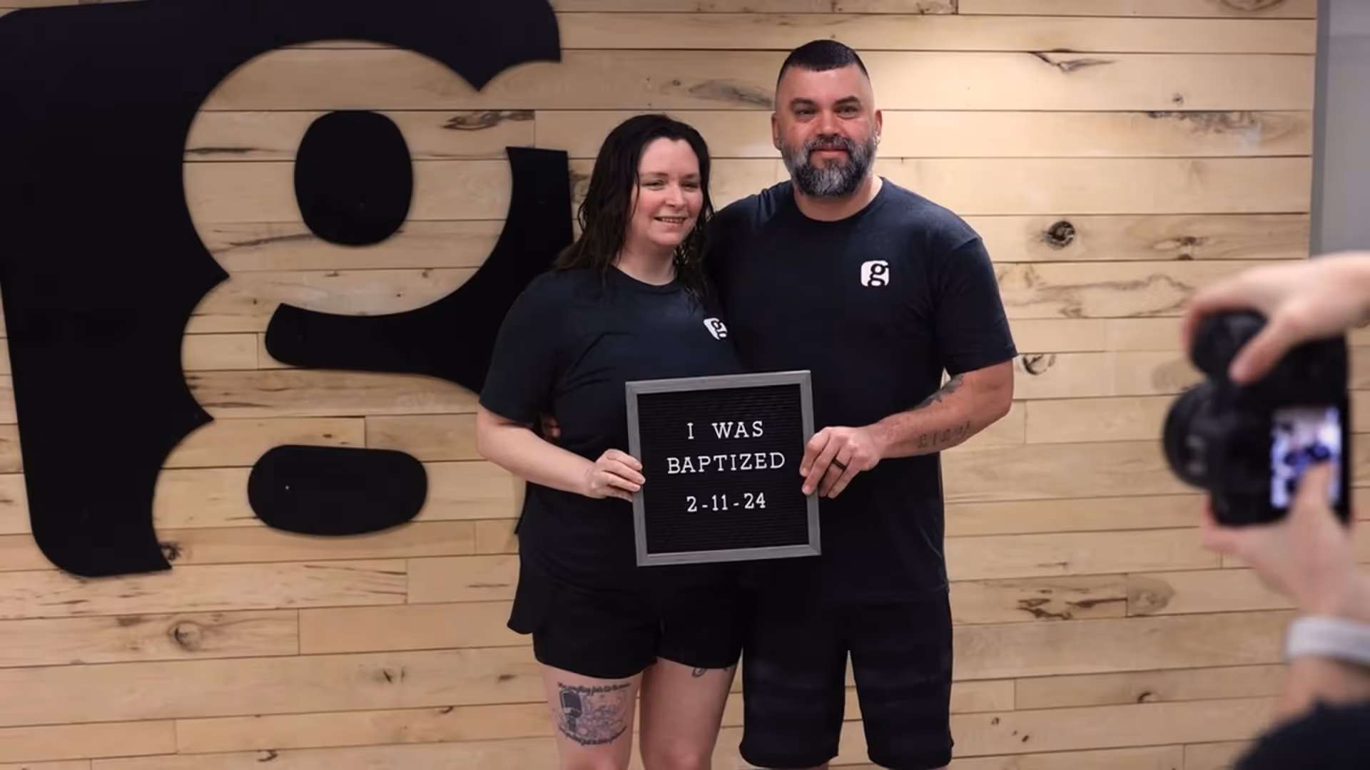 Couple smiling while holding a baptism sign in front of a wooden wall with a large “g” logo.