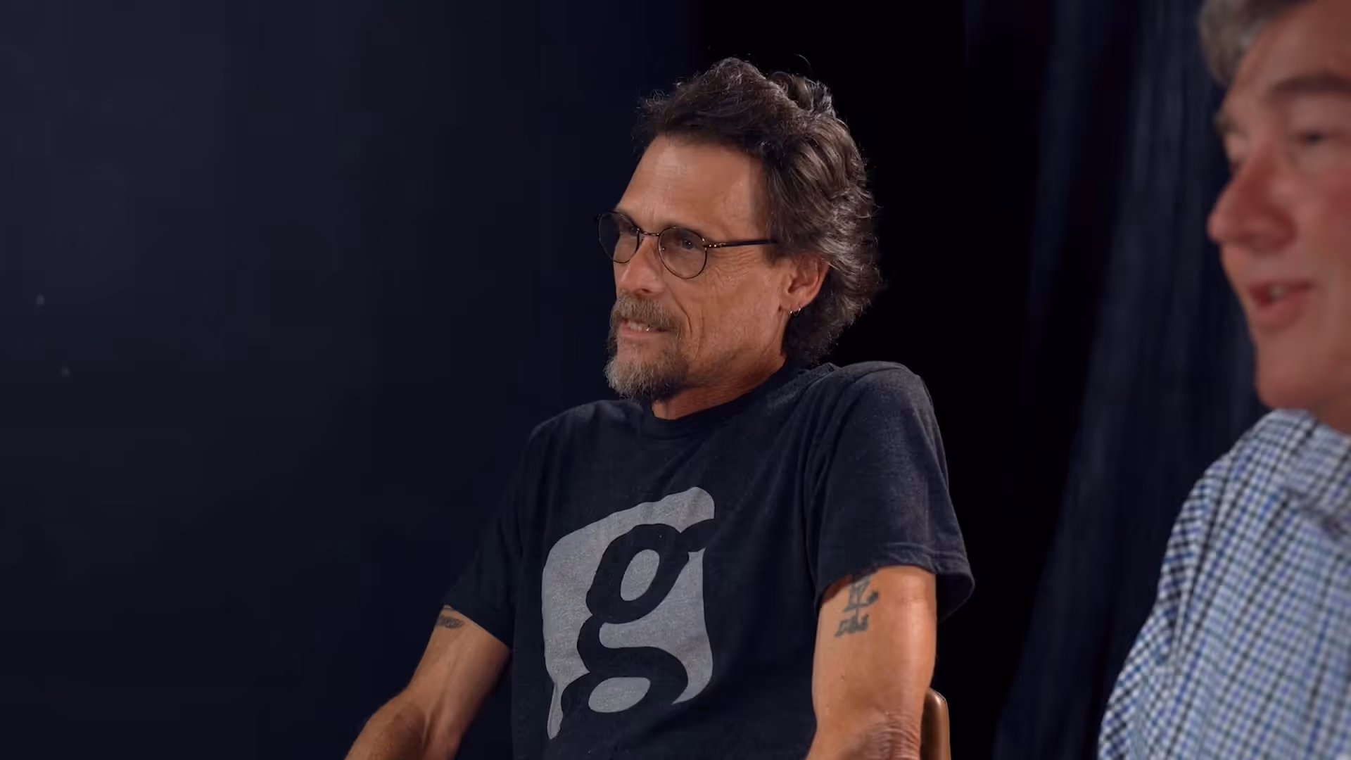Man with glasses speaking in a dim studio setting with another person blurred beside him.