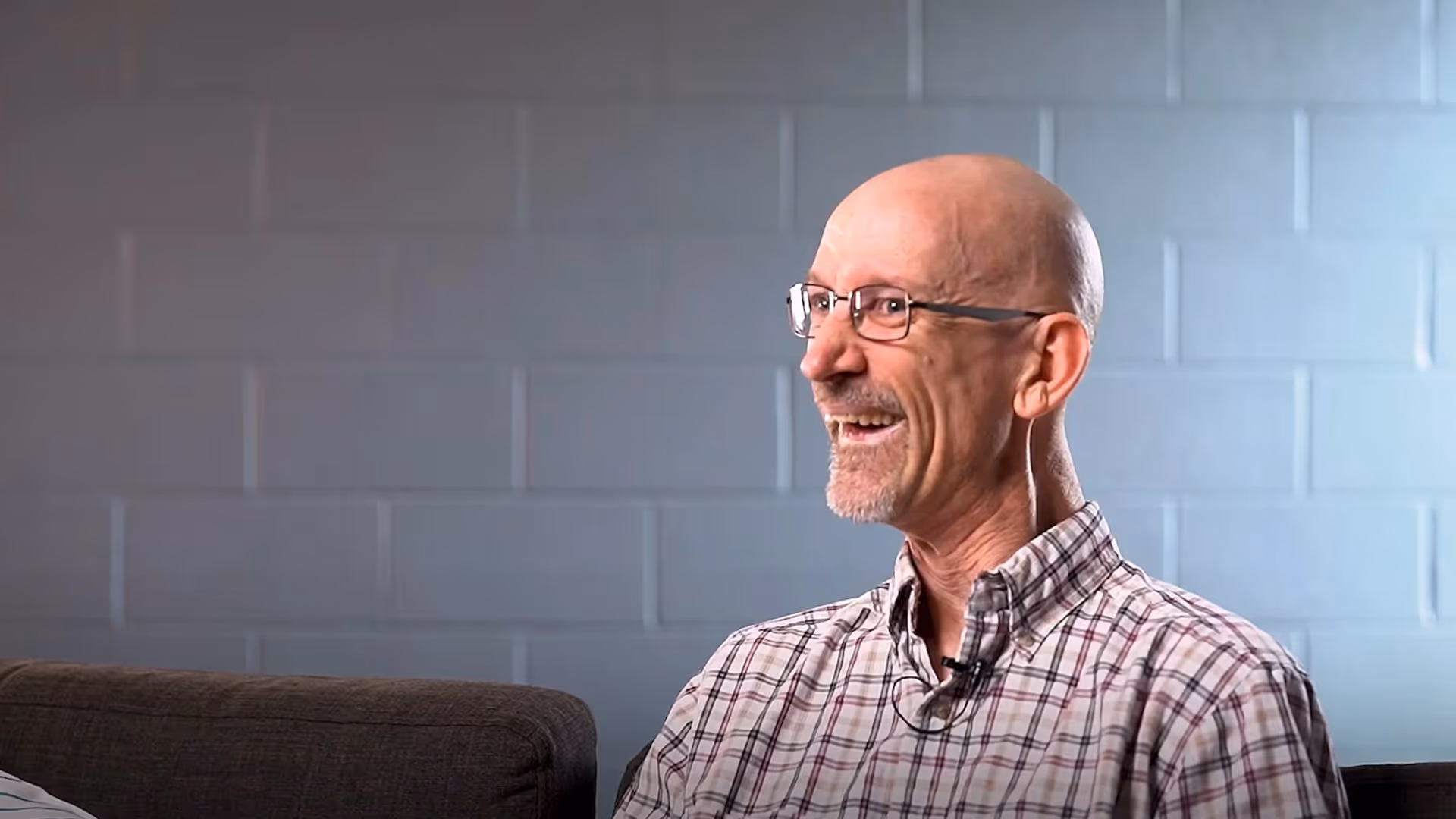 Smiling man with glasses seated in a softly lit room with a pale brick wall.