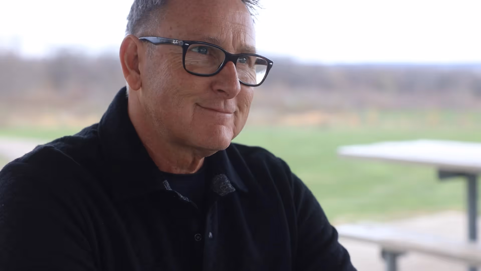 Smiling man with glasses sitting outdoors with fields in the background.