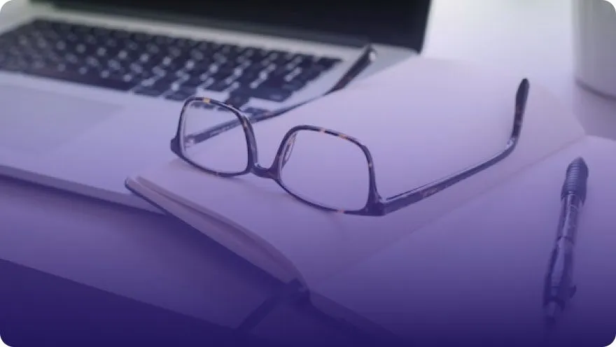 Reading glasses resting on an open notebook next to a laptop and pen on a desk