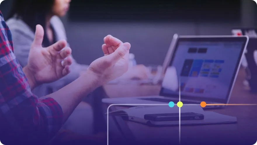 Person gesturing while discussing near a laptop showing colorful app designs, purple overlay