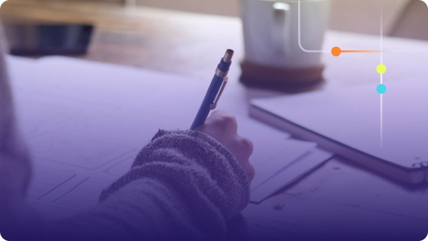 Hand holding a pen writing on paper at a desk with a coffee mug and tablet nearby