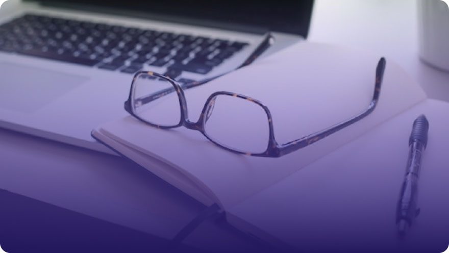 Reading glasses resting on an open notebook next to a laptop and pen on a desk