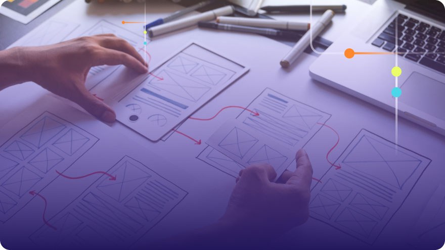 Hands arranging UX wireframe sketches on a desk with a laptop and markers nearby