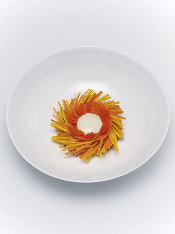 Création culinaire graphique en forme de fleur orangée, servie dans une assiette creuse blanche immaculée.