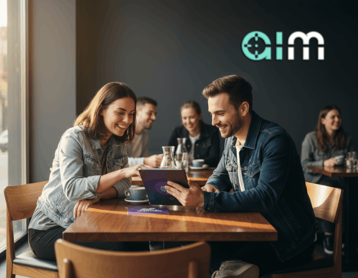 Two young adults sitting at a wooden table in a cafe, smiling and looking at a tablet together with coffee cups nearby.