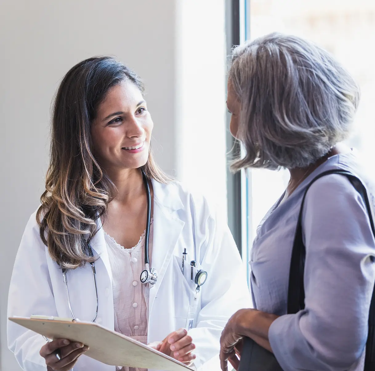 Smiling female doctor with stethoscope talking to a woman by a window, holding a clipboard.
