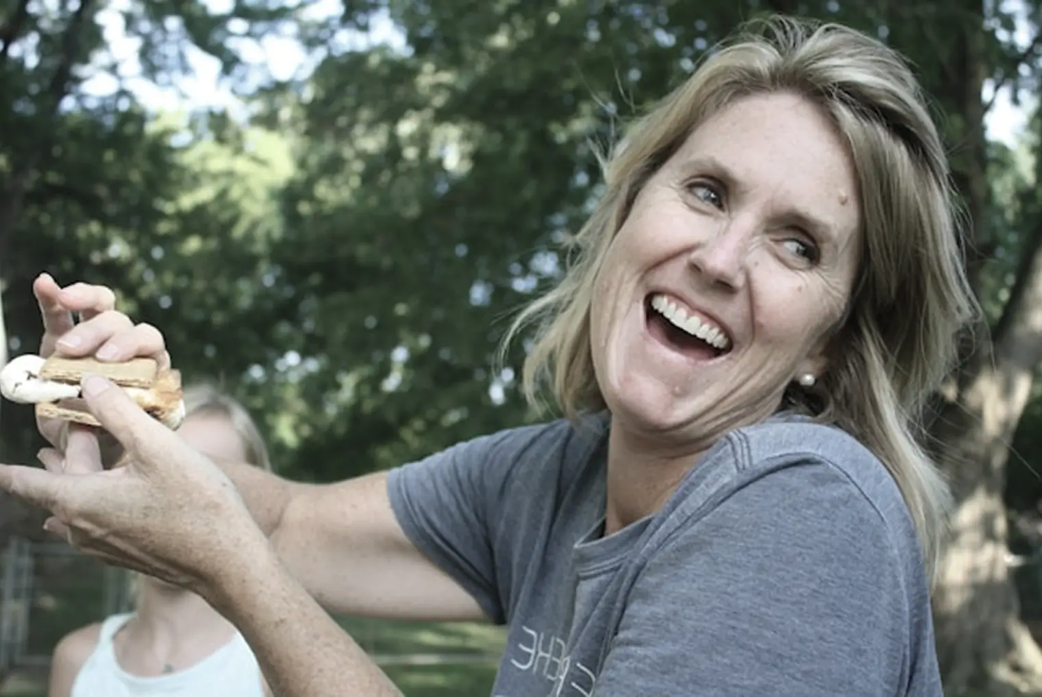 Smiling woman holding a s'more outdoors with trees in the background.