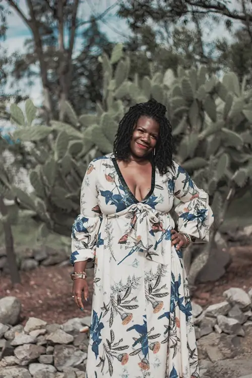 Smiling woman with braided hair wearing a white floral dress standing outdoors in front of large cactus plants.