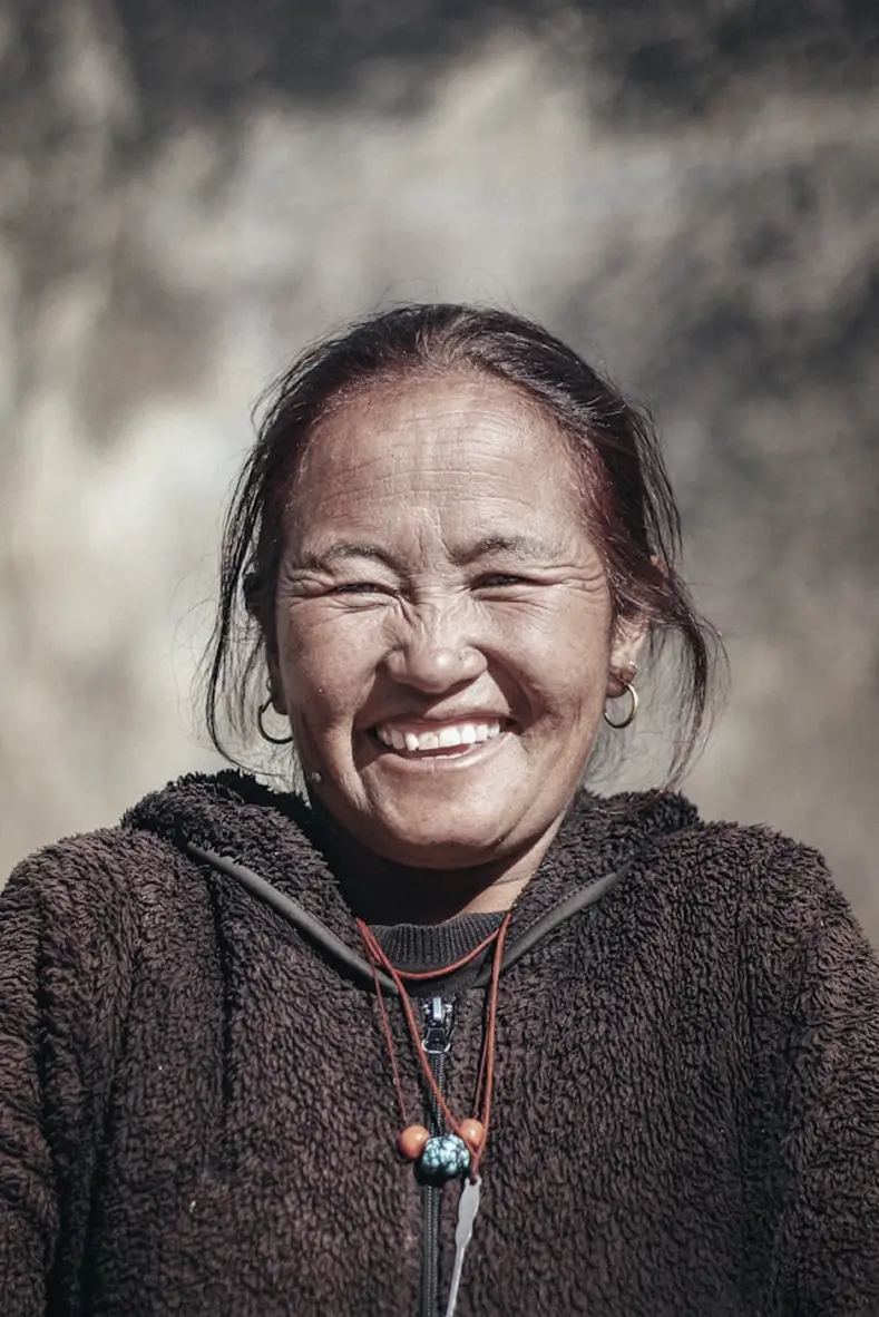 Smiling elderly woman wearing a dark fuzzy jacket and beaded necklace against a blurred background.
