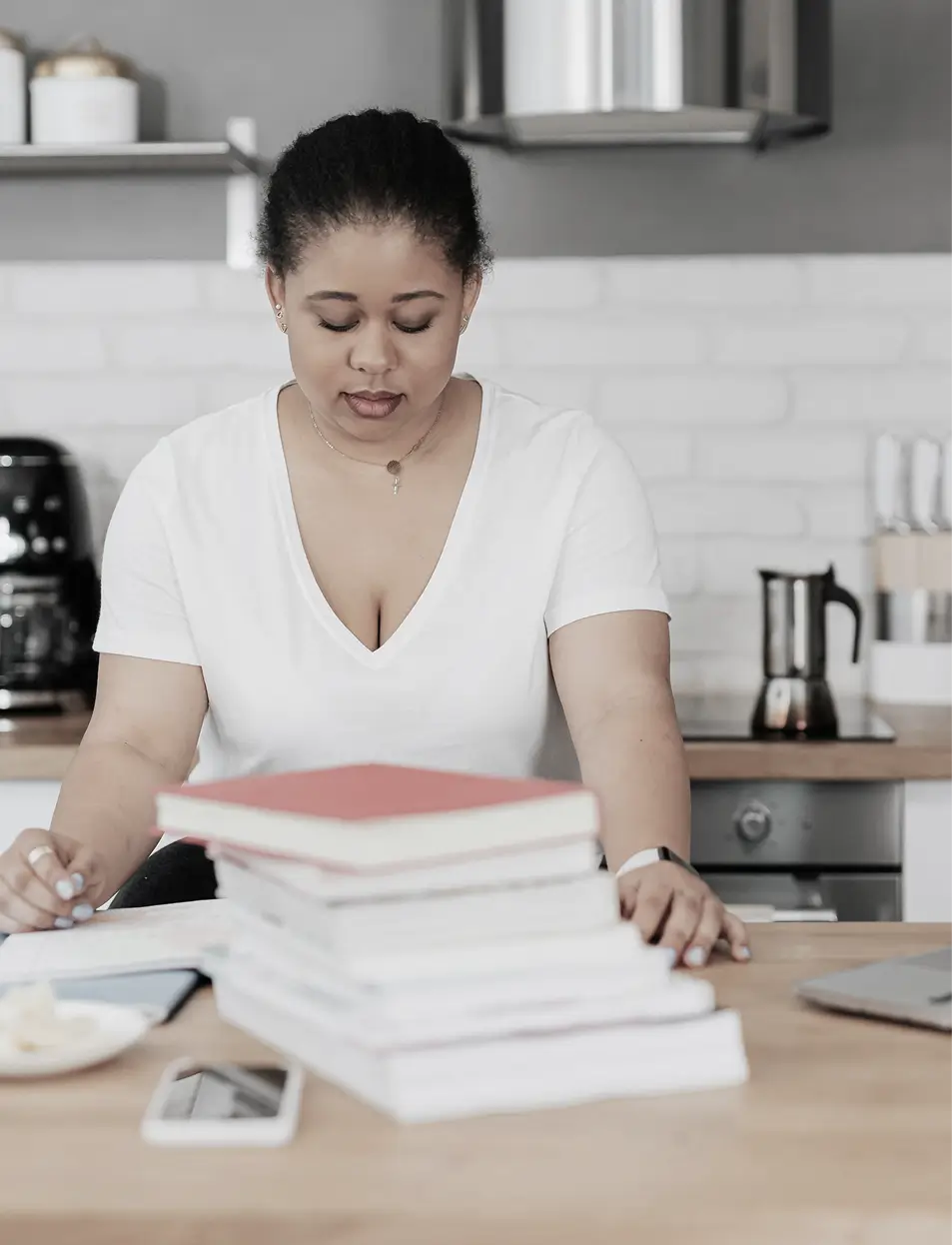 Woman in a white shirt sitting at a wooden table with a stack of books, a smartphone, and a plate with food in a kitchen.