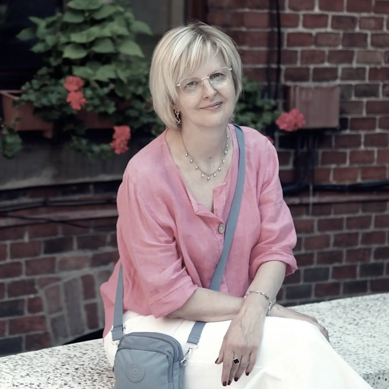 Smiling woman with short blonde hair, glasses, and a pink blouse sitting outdoors with a gray crossbody bag and brick wall with plants in the background.