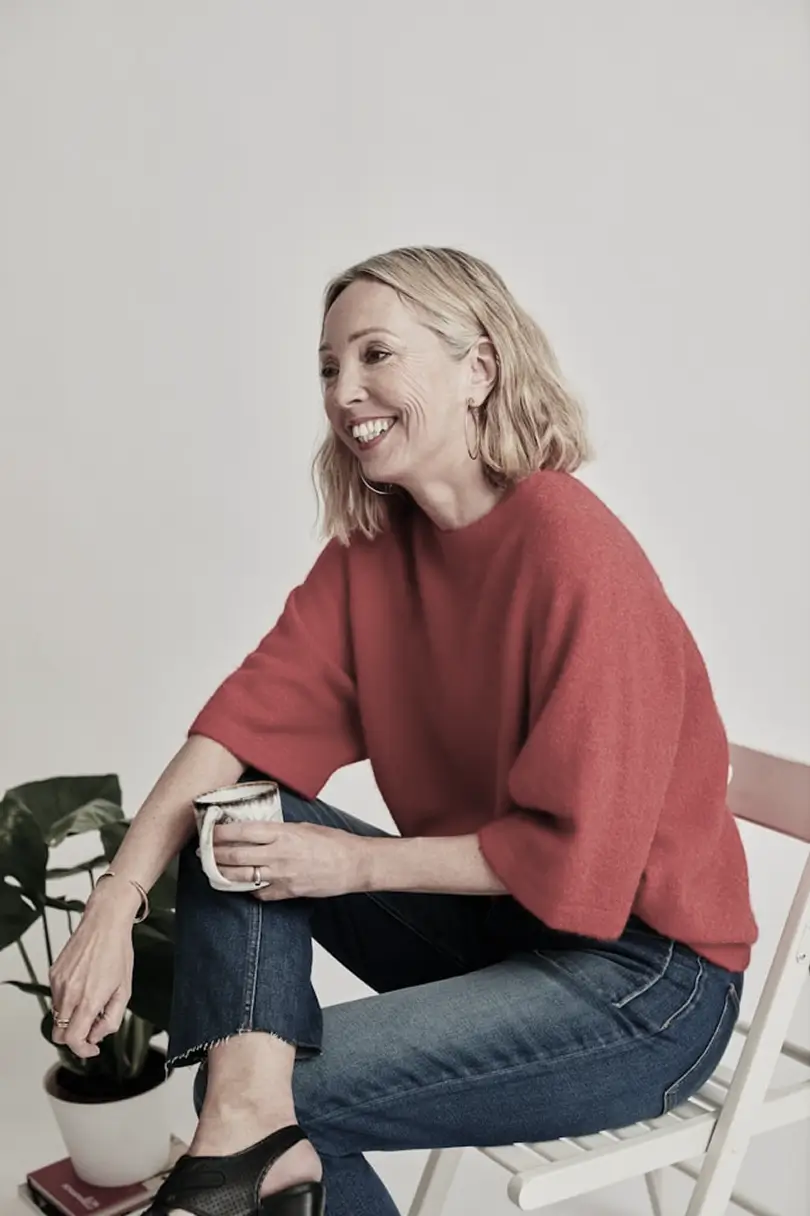 Smiling woman with blonde hair wearing a red sweater and blue jeans, sitting on a white chair holding a mug.