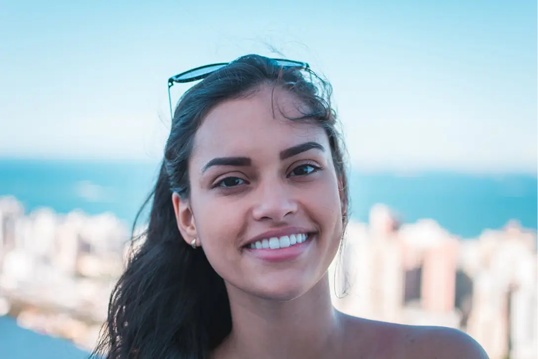 Close-up of a smiling young woman with long dark hair, sunglasses on her head, with a blurred cityscape and blue sky in the background.